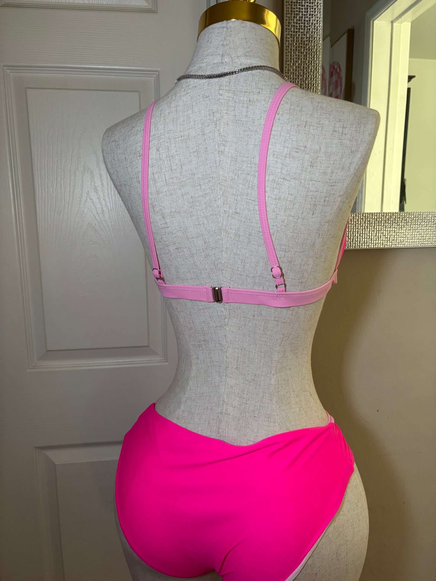 A gray fabric dress form mannequin torso wearing a bright pink bralette with thin adjustable straps and a small center clasp, photographed indoors against a white paneled door.