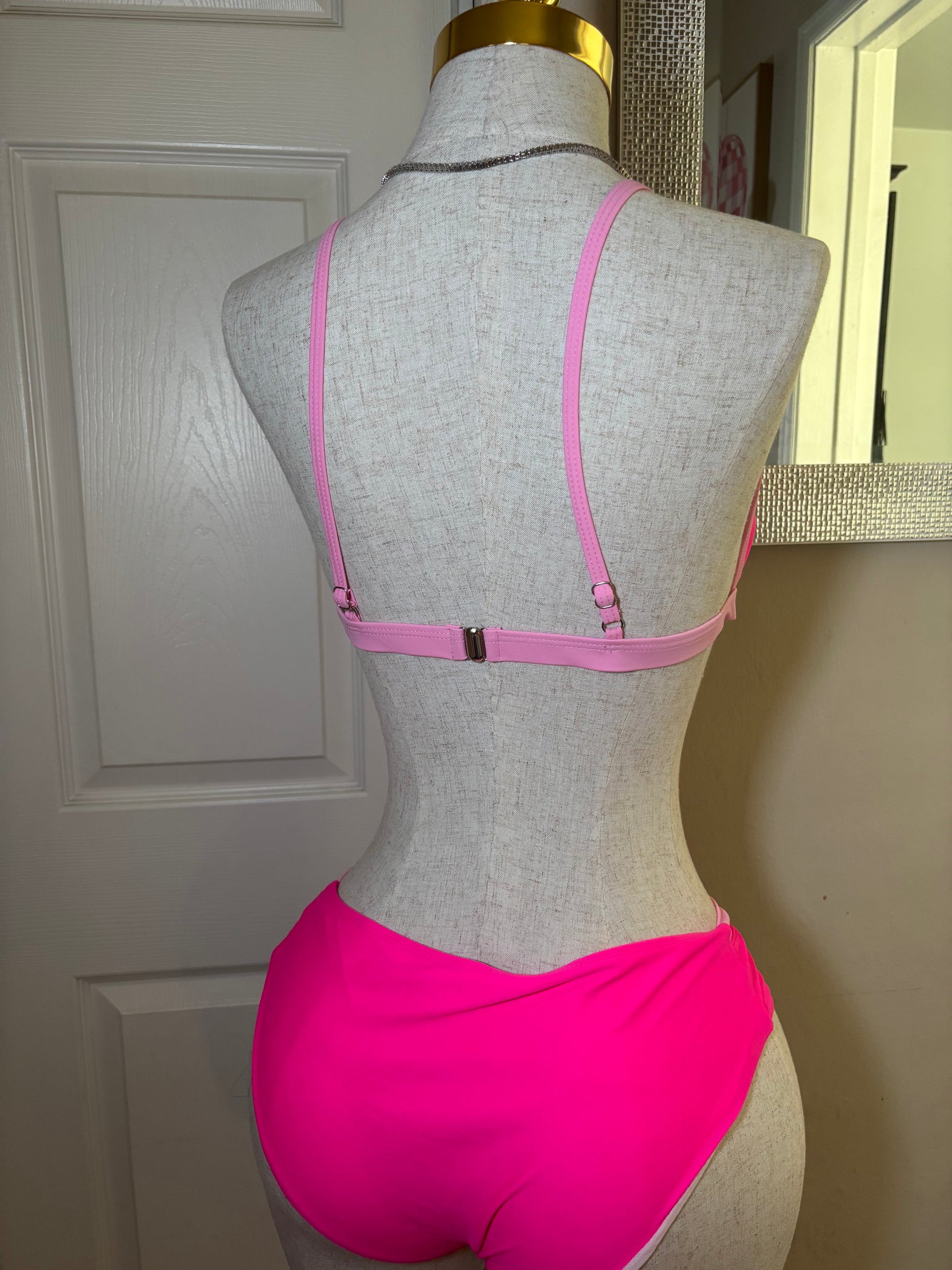 A gray fabric dress form mannequin torso wearing a bright pink bralette with thin adjustable straps and a small center clasp, photographed indoors against a white paneled door.